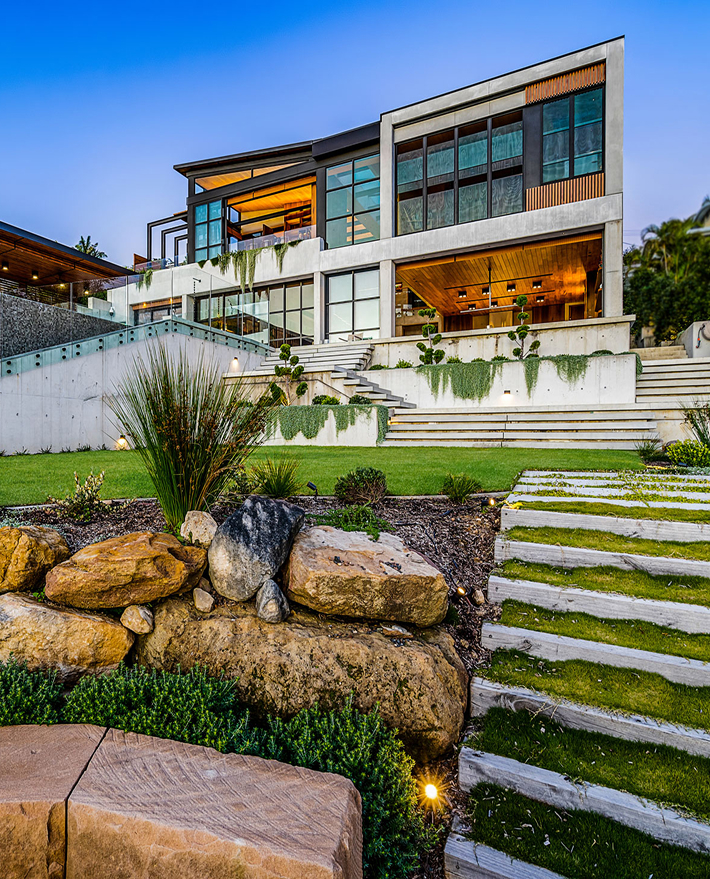 Dramatic lower view of terraced landscape with sandstone and spiky greenery, with the main house rising up at the back; characterised by polished concrete and floor to ceiling windows