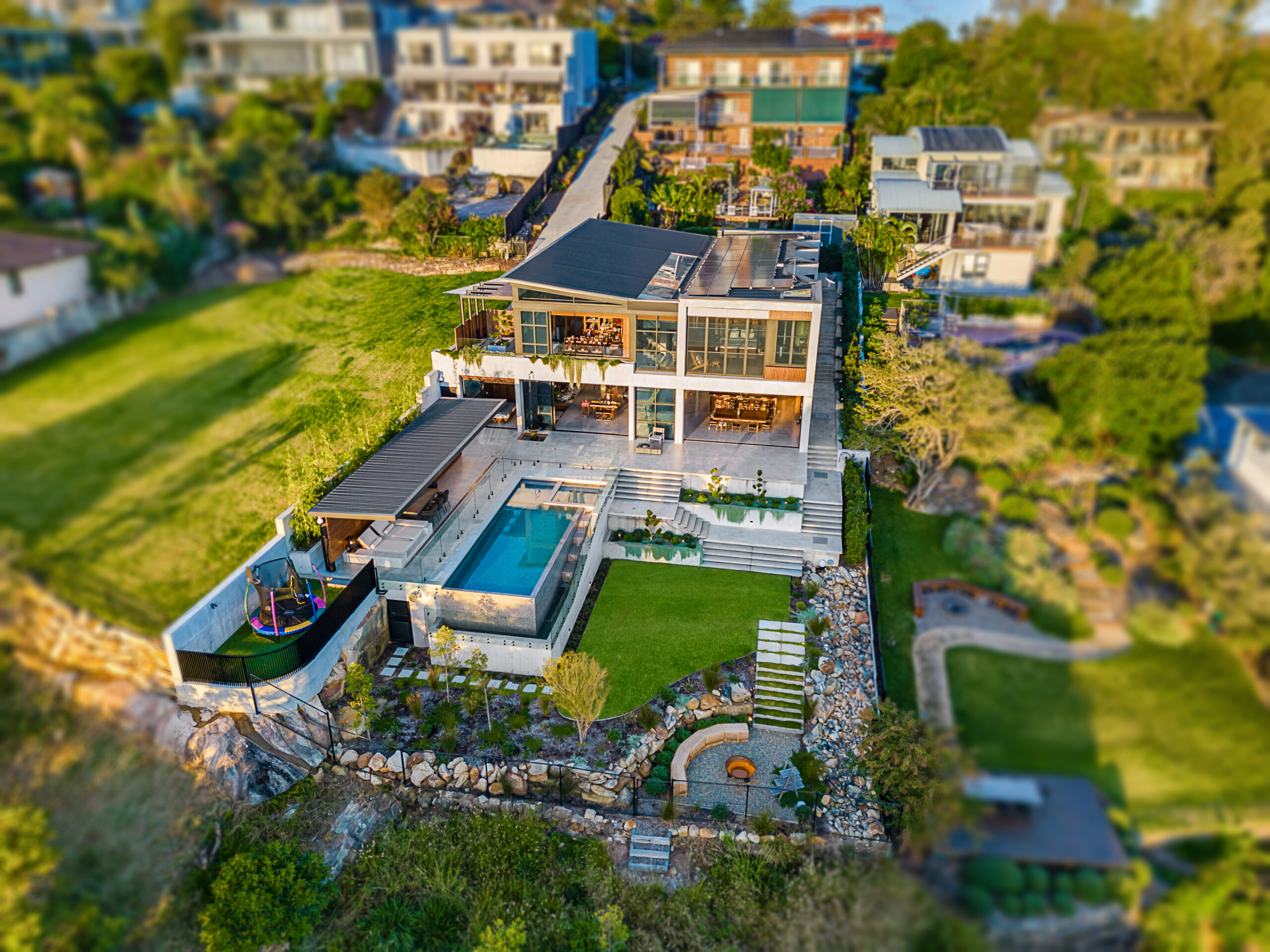 Elevated shot of expansive, ultra-modern house featuring charcoal accents and polished concrete from the backyard up to the house. The terraced backyard includes a firepit space, blue pool and lounging deck, trampoline and foliage draping across the terraced levels.