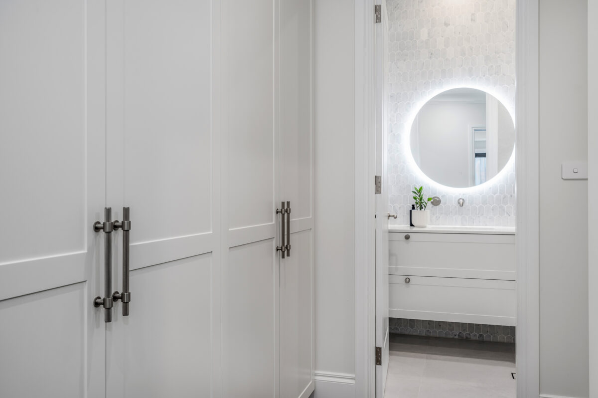 Ensuite bathroom viewed from wardrobe area, with white cabinetry, round illuminated mirror and feature wall tiles