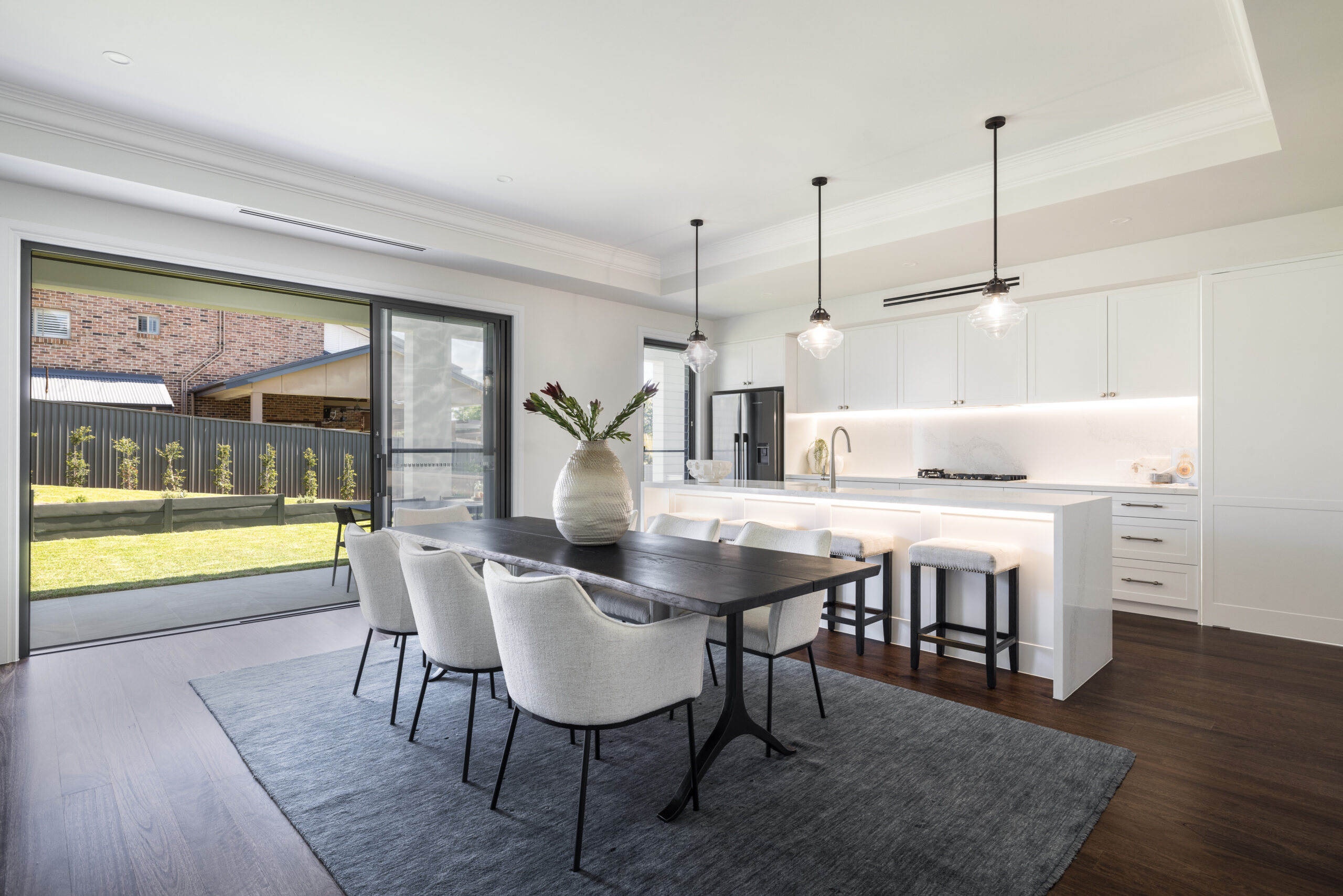 Open-plan kitchen and dining area with white cabinetry, pendant lights, timber flooring and sliding doors to the backyard