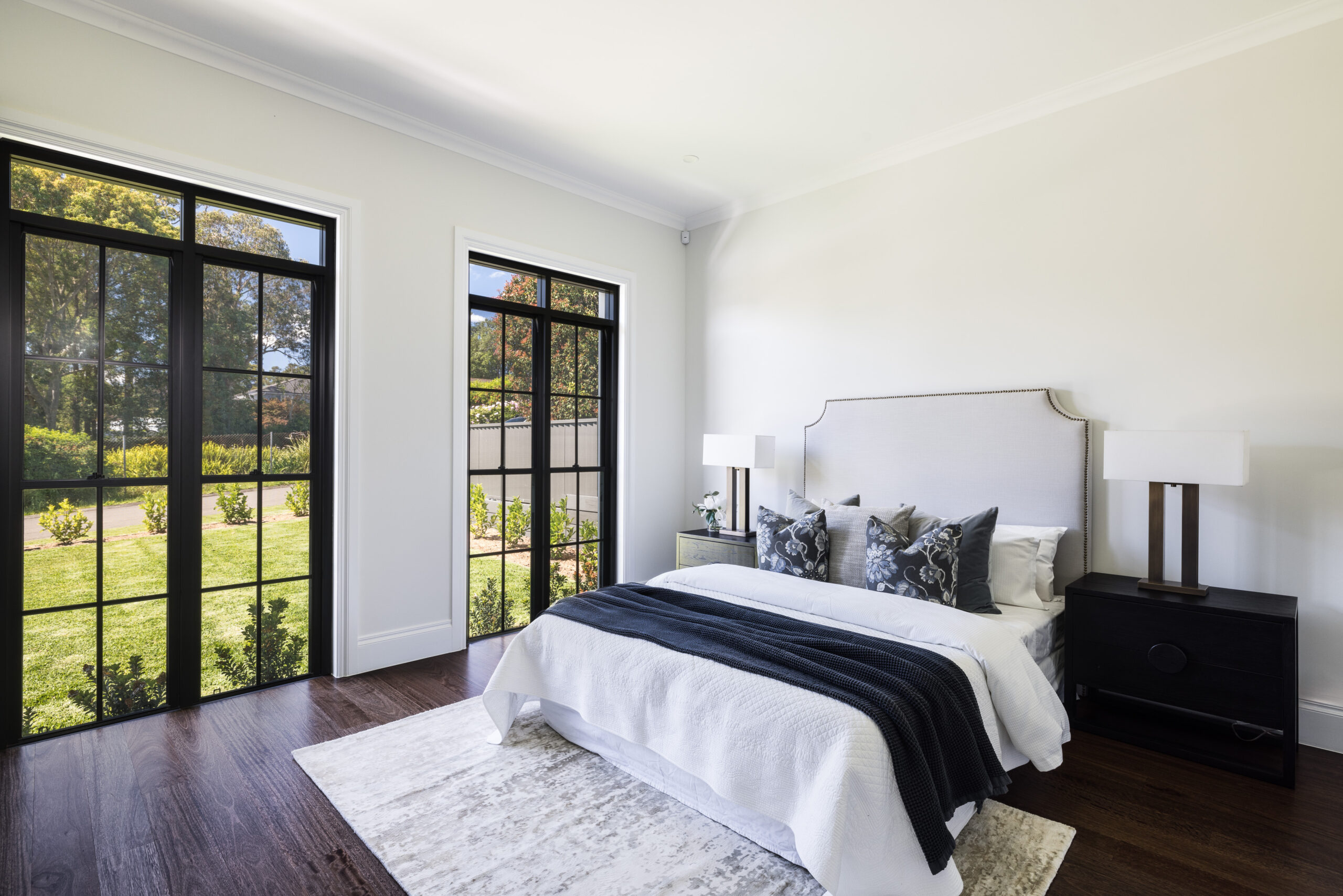 Light-filled bedroom with upholstered bed, dark timber flooring and black-framed glass doors overlooking the garden