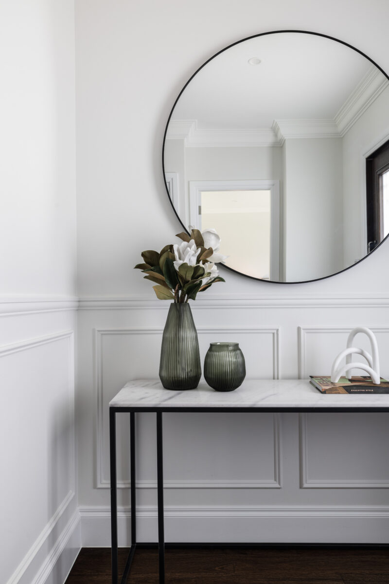 Entryway console with round mirror, marble tabletop and decorative vases against white wall panelling